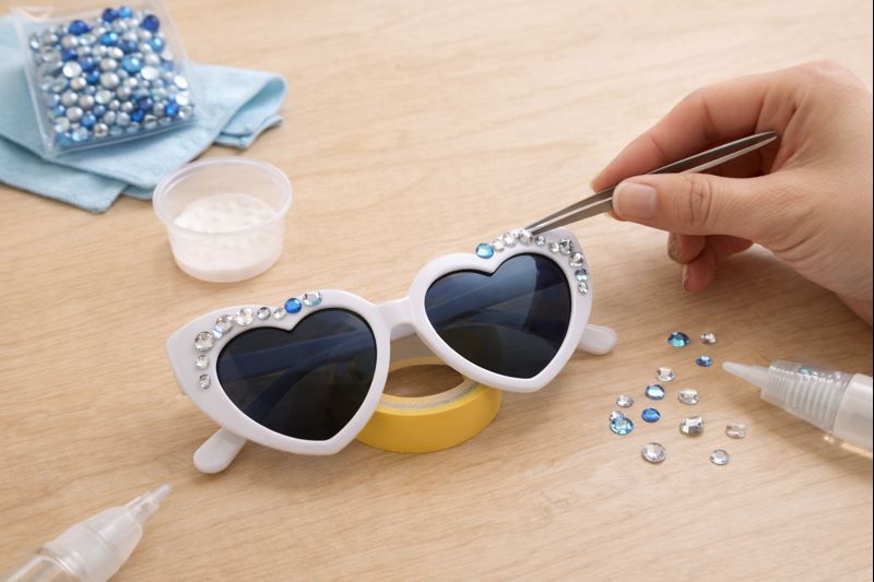 手元の作業工程が分かる写真風イメージ。サングラスフレームにラインストーンを仮置きしている場面、マスキングテープ、ピンセット、少量の接着剤、乾燥中に浮かせて置く工夫が分かる小道具。顔は写さない。作業台は明るい木目、清潔感。
