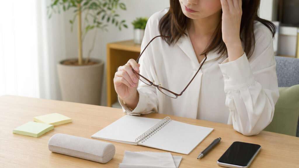 乱視メガネの違和感でこめかみに手を当て、メガネを持つ女性の手元（室内のテーブル）